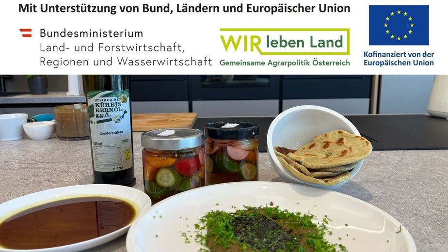 Auf einer Küchentheke sind die zubereiteten Gerichte angerichtet: Ein Teller mit Käferbohnen-Hummus, garniert mit frischen Kräutern, eine Schale mit Kernöl-Flatbread, zwei Gläser mit sauer eingelegtem Gemüse und eine Flasche steirisches Kürbiskernöl.