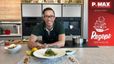 Koch Peter Lehner präsentiert lächelnd in einer modernen Küche den fertig angerichteten Kabeljau mit Riesling Beurre Blanc. Das Gericht ist auf einem weißen Teller angerichtet und mit Zuckerschotensalat sowie Croutons garniert, umgeben von weiteren frischen Zutaten auf der Kücheninsel.