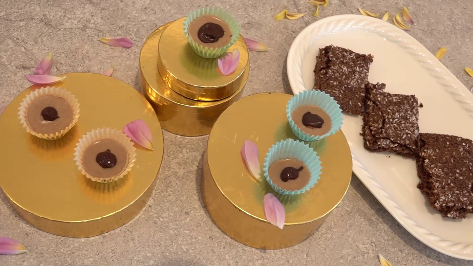 Hausgemachte Toffees in bunten Papierförmchen auf goldenen Tellern, jeweils mit einer Haselnuss und einem Schokoladenpunkt verziert, neben einem Teller mit geschnittenen veganen Kokosnuss-Brownies. Rosa Blütenblätter sind dekorativ um die Süßigkeiten verstreut.