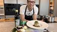 Chef Peter Lehner präsentiert lächelnd den fertigen Käferbohnenknödel mit Schmelzzwiebeln auf einem weißen Teller. Im Vordergrund sind die regionalen Hauptzutaten wie steirisches Kürbiskernöl, eine Dose Käferbohnen, Zwiebeln und Kürbiskerne dekorativ angerichtet.