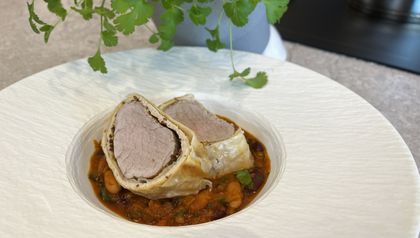 Zwei Scheiben Schweinsfilet im goldbraunen Strudelteig, angerichtet auf einem herzhaften Bohnen-Cassoulet mit weißen und Kidneybohnen. Das Gericht wird in einer weißen, strukturierten Schüssel serviert, im Hintergrund ist eine grüne Kräuterpflanze erkennbar.