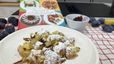 Ein Teller mit frisch zubereitetem, fluffigem Kaiserschmarrn, der großzügig mit Staubzucker bestreut ist. Im Hintergrund stehen eine kleine Schale mit Zwetschkenröster, frische Zwetschken und das Kochbuch „Kochen mit Dampf & Leidenschaft“ auf einem rot-weiß karierten Küchentuch.
