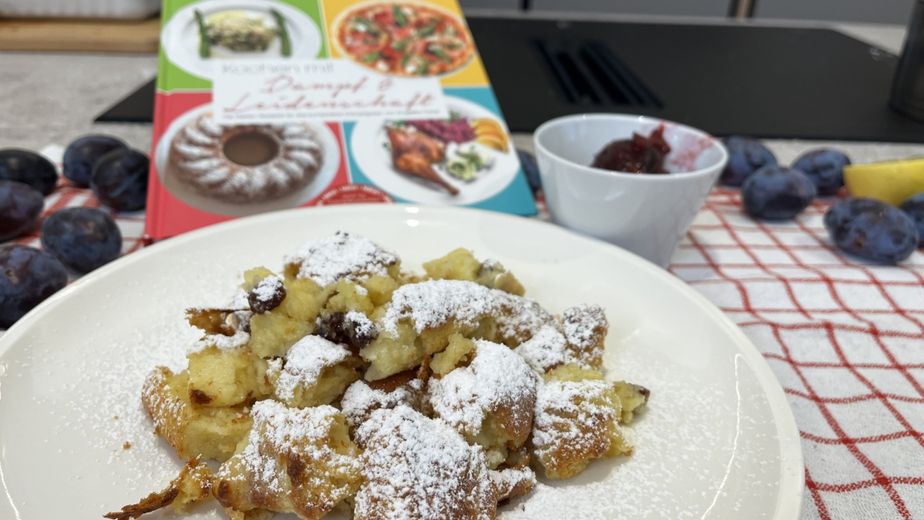 Ein Teller mit frisch zubereitetem, fluffigem Kaiserschmarrn, der großzügig mit Staubzucker bestreut ist. Im Hintergrund stehen eine kleine Schale mit Zwetschkenröster, frische Zwetschken und das Kochbuch „Kochen mit Dampf & Leidenschaft“ auf einem rot-weiß karierten Küchentuch.