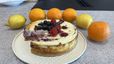 Angeschnittene Topfentorte mit Himbeer-Swirl, garniert mit frischen Blaubeeren und Himbeeren auf einem weißen Teller. Die Schnittkante offenbart die Marmorierung durch die Himbeermasse im Inneren des Kuchens. Zitronen und Orangen liegen dekorativ daneben.