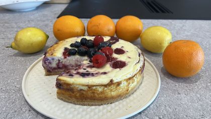 Angeschnittene Topfentorte mit Himbeer-Swirl, garniert mit frischen Blaubeeren und Himbeeren auf einem weißen Teller. Die Schnittkante offenbart die Marmorierung durch die Himbeermasse im Inneren des Kuchens. Zitronen und Orangen liegen dekorativ daneben.