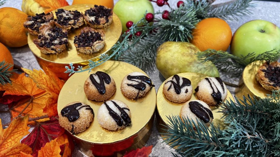 Die fertig gebackenen veganen Florentiner und Kokosmakronen sind auf goldenen Podesten angerichtet. Die runden Kokosbällchen und die flachen, nussigen Florentiner sind jeweils mit dunklen Schokoladenstreifen verziert. Das Gebäck ist festlich zwischen Tannenzweigen, Äpfeln und Orangen dekoriert.