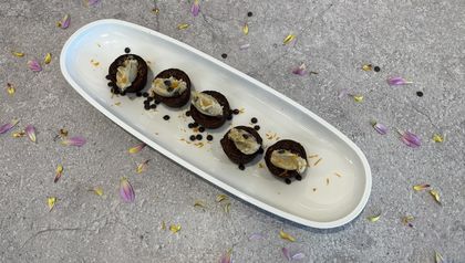 Fünf fertige Brownies sind auf einer länglichen, weißen Servierplatte angerichtet. Auf jedem Brownie sitzt eine Nocke aus Cookie Dough, die mit Schokotropfen bestreut ist. Weitere Schokotropfen und dekorative Blütenblätter sind um die Brownies auf der Platte und dem grauen Untergrund verteilt.