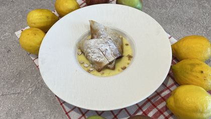 Zwei Stücke goldbrauner Mini-Apfelstrudel, mit Puderzucker bestäubt, serviert in einem tiefen weißen Teller mit cremiger Vanillesauce und gehackten Walnüssen. Der Teller ist auf einem Küchentuch angerichtet und von frischen Äpfeln und Zitronen umgeben, die die Hauptzutaten des Rezepts widerspiegeln.