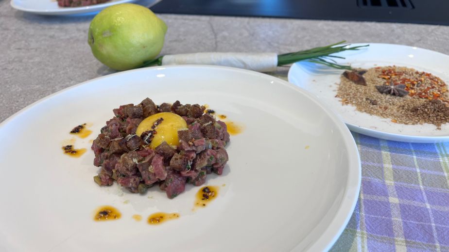 Ein weißer Teller mit handgeschnittenem Beef Tatar, das mit einem gebeizten Eigelb und knusprigen Croutons angerichtet ist. Um das Fleisch herum sind feine Tropfen Chili-Öl verteilt, während im Hintergrund frische Zutaten wie eine Zitrone, Schnittlauch und Gewürze bereitliegen.