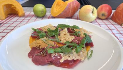 Ein weißer Teller mit fein aufgeschnittenem Rinder-Carpaccio auf einer cremigen, orangefarbenen Kürbiscreme. Das Gericht ist garniert mit knusprigen Parmesanchips, frischen grünen Kräutern und etwas Chili-Öl. Im leicht verschwommenen Hintergrund liegen herbstliche Zutaten wie Kürbisse und Äpfel auf einem rot-weiß karierten Tuch.