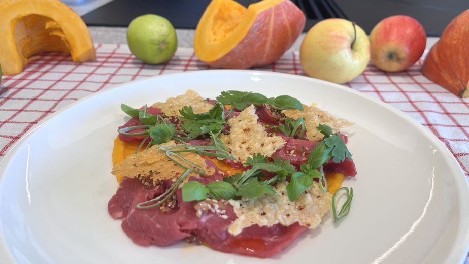 Ein weißer Teller mit fein aufgeschnittenem Rinder-Carpaccio auf einer cremigen, orangefarbenen Kürbiscreme. Das Gericht ist garniert mit knusprigen Parmesanchips, frischen grünen Kräutern und etwas Chili-Öl. Im leicht verschwommenen Hintergrund liegen herbstliche Zutaten wie Kürbisse und Äpfel auf einem rot-weiß karierten Tuch.