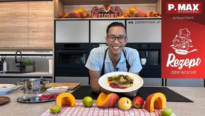 Chefkoch Peter Lehner präsentiert lächelnd einen Teller mit dem zubereiteten Carpaccio in einer modernen Showküche. Im Vordergrund sind herbstliche Zutaten wie halbierte Kürbisse, Äpfel und Limetten auf der Arbeitsplatte angerichtet.