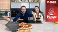 Max Impertro und Tanja lächeln in einer modernen Küche hinter einer frisch gebackenen Salami-Pizza mit Basilikum auf einem Holzbrett. Max hält eine graue Pizzaschaufel in der Hand, während Tanja ein einzelnes Basilikumblatt präsentiert.
