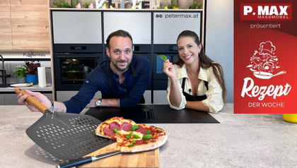 Max Impertro und Tanja lächeln in einer modernen Küche hinter einer frisch gebackenen Salami-Pizza mit Basilikum auf einem Holzbrett. Max hält eine graue Pizzaschaufel in der Hand, während Tanja ein einzelnes Basilikumblatt präsentiert.